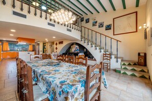 Uma sala de jantar com uma mesa comprida, cadeiras, um lustre, uma escadaria e uma lareira.
