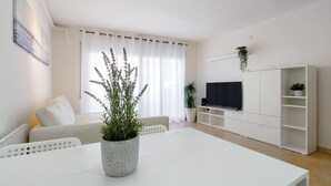 Una sala de estar moderna con un sofá blanco, una mesa de comedor, un televisor sobre un mueble blanco y una planta en maceta.