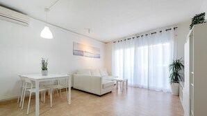 Una sala de estar moderna con un sofá blanco, una mesa de comedor y sillas, una planta en maceta y un gran ventanal con cortinas traslúcidas.