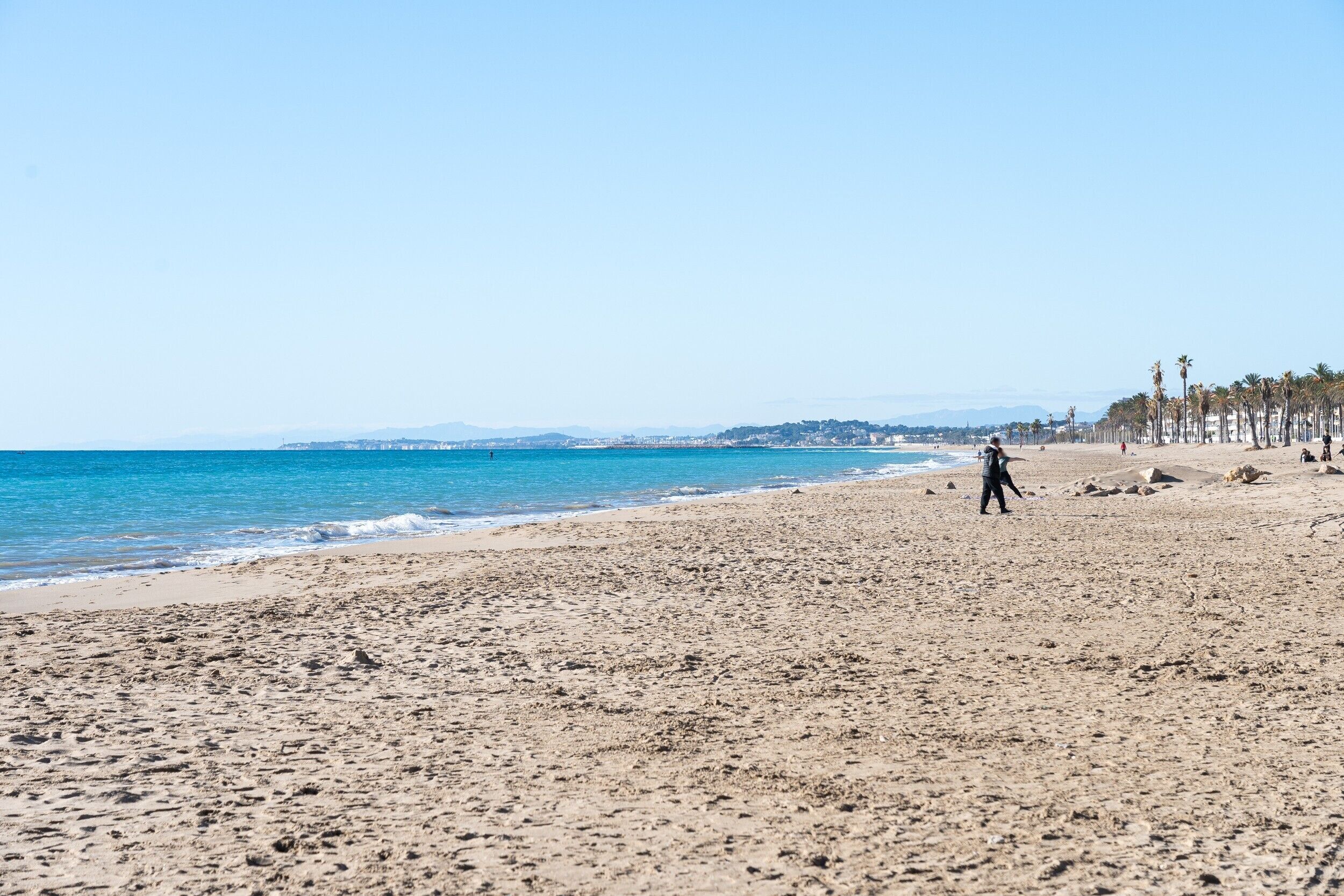 A beach with a person walking, clear skies, and a distant cityscape.