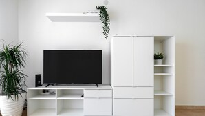 Un salón moderno con televisor de pantalla plana, muebles blancos, una planta en maceta y un cartel con la palabra 'Welcome'.
