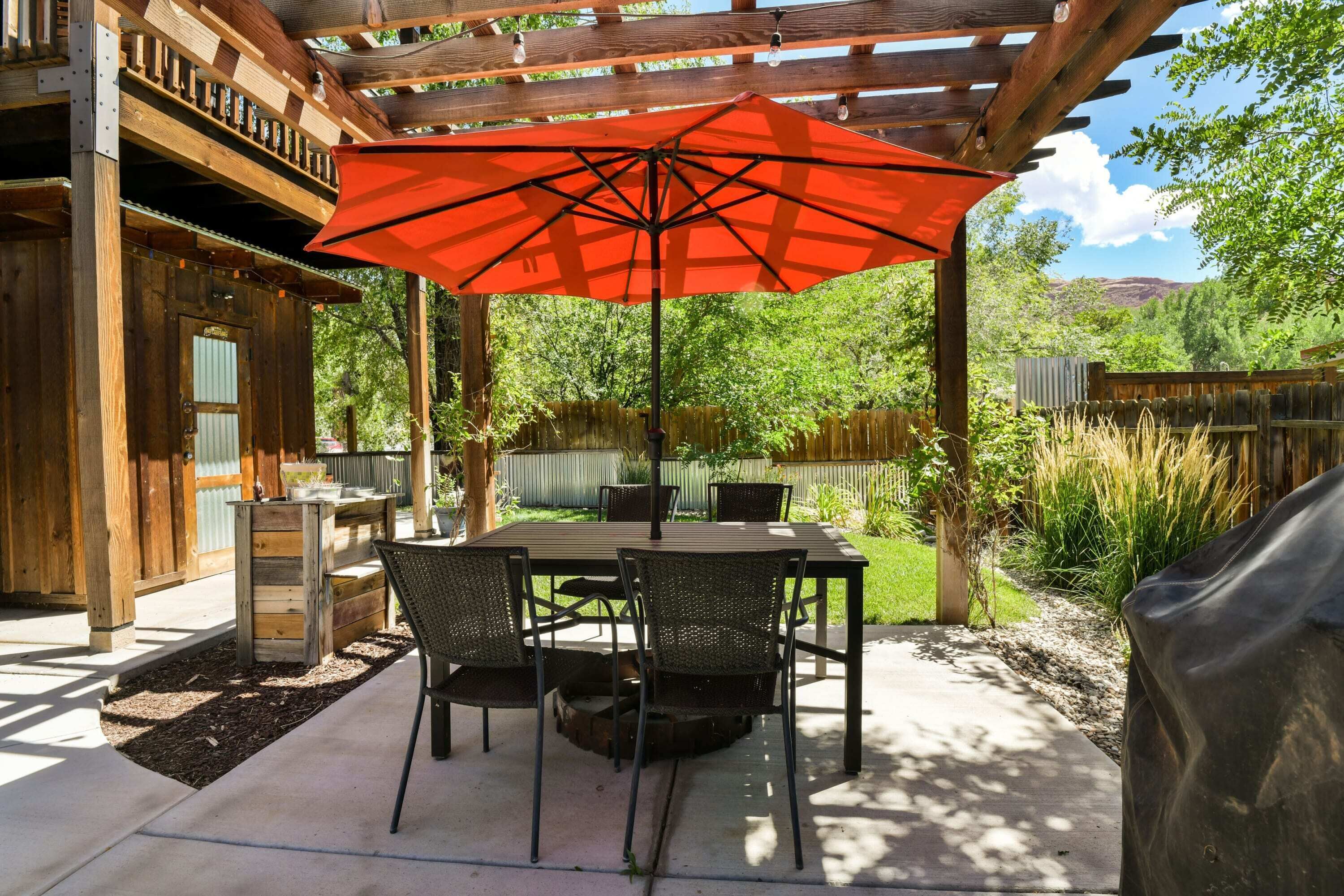 An outdoor patio with a wooden pergola, a table with chairs, and a red umbrella.