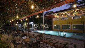 Un espace extérieur avec une piscine, dont le sol est recouvert de carreaux, entouré de chaises et de tables, et un bâtiment jaune doté de balcons.