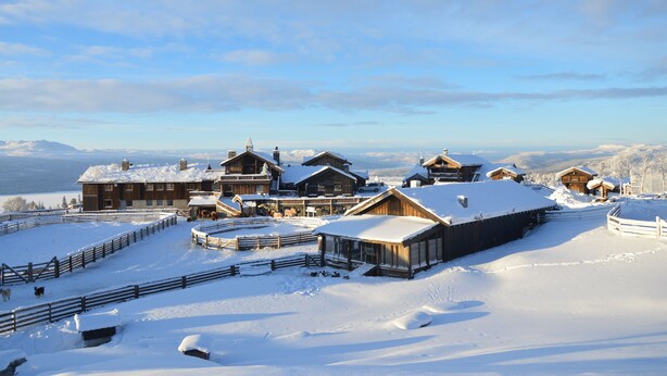 Eine schneebedeckte Lodge mit mehreren Holzhütten, einem eingezäunten Hof und freiem Blick auf die umliegenden Berge.