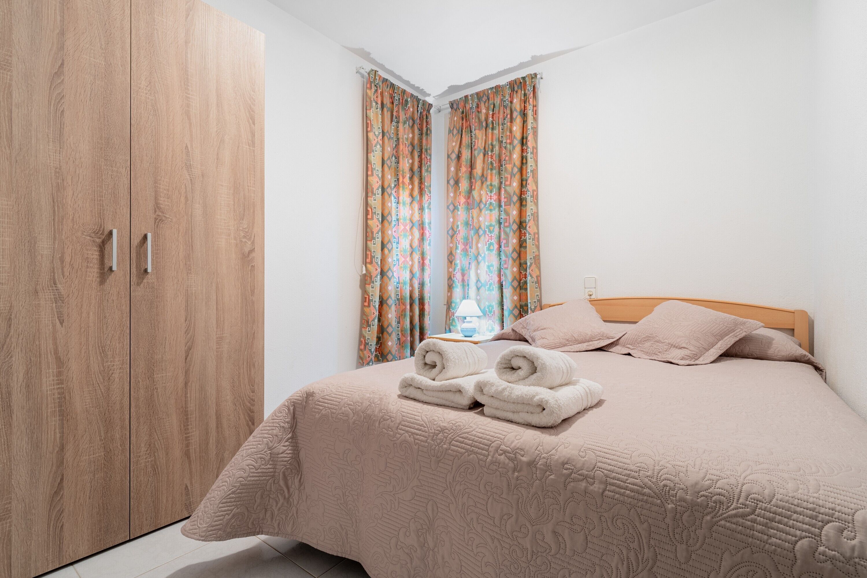 Een slaapkamer met een houten garderobe, een bed met een beige deken en kamergordijnen met een patroon.