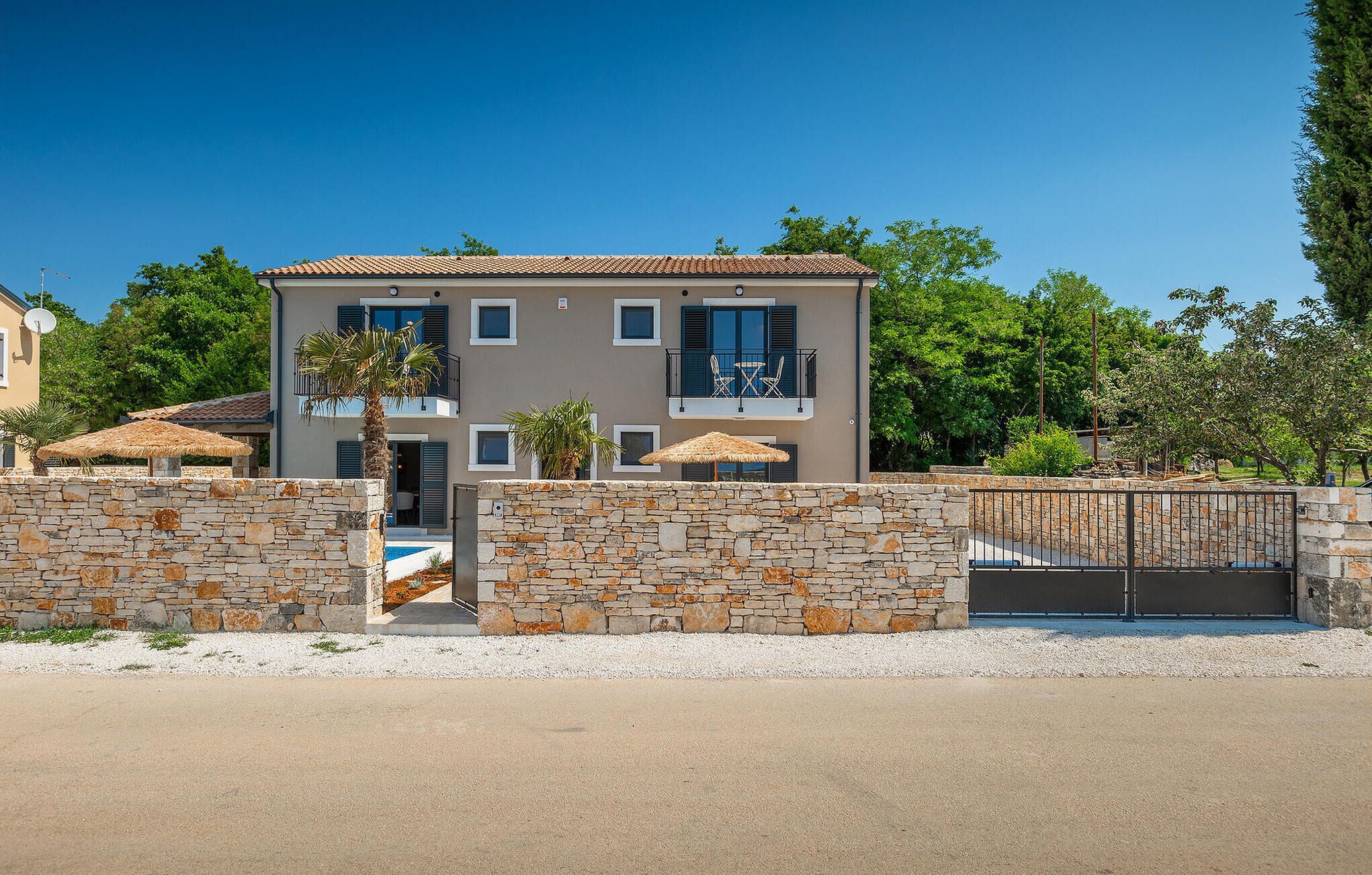 A modern two-story house with a stone wall entrance, a balcony with outdoor furniture, and a clear blue sky.