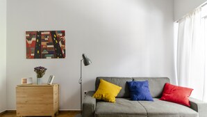 A living room with a grey sofa, wooden side table, floor lamp, and a colorful abstract painting on the wall. - Colourful Apartment Under the Cropolis (Athens)