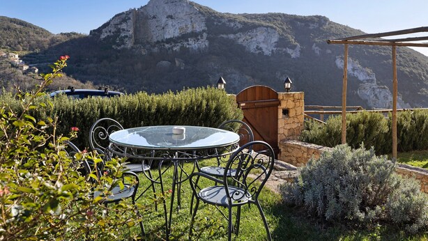 A round glass table with metal chairs, a stone wall, and a wooden pergola in a garden with a mountainous backdrop. - Apartment "Casa Virginia - Corbezzolo" with Shared Pool & Wi-Fi (Orco Feglino)