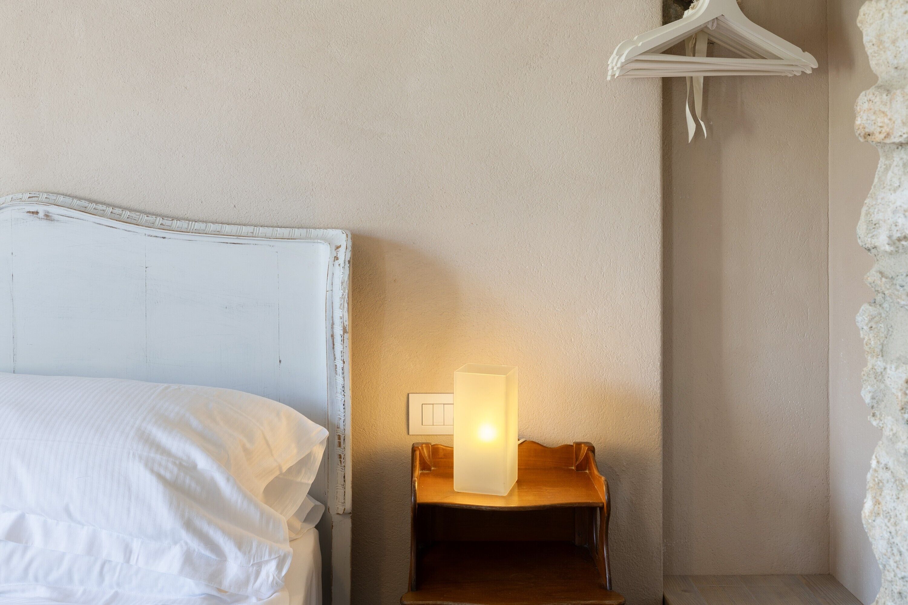 A bed with a white headboard, a bedside table with a lamp, and a wall-mounted light switch.