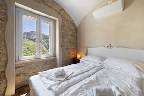 Un dormitorio con pared de piedra, cama con ropa blanca, mesita de noche con lámpara y ventana con vistas a vegetación y un acantilado.