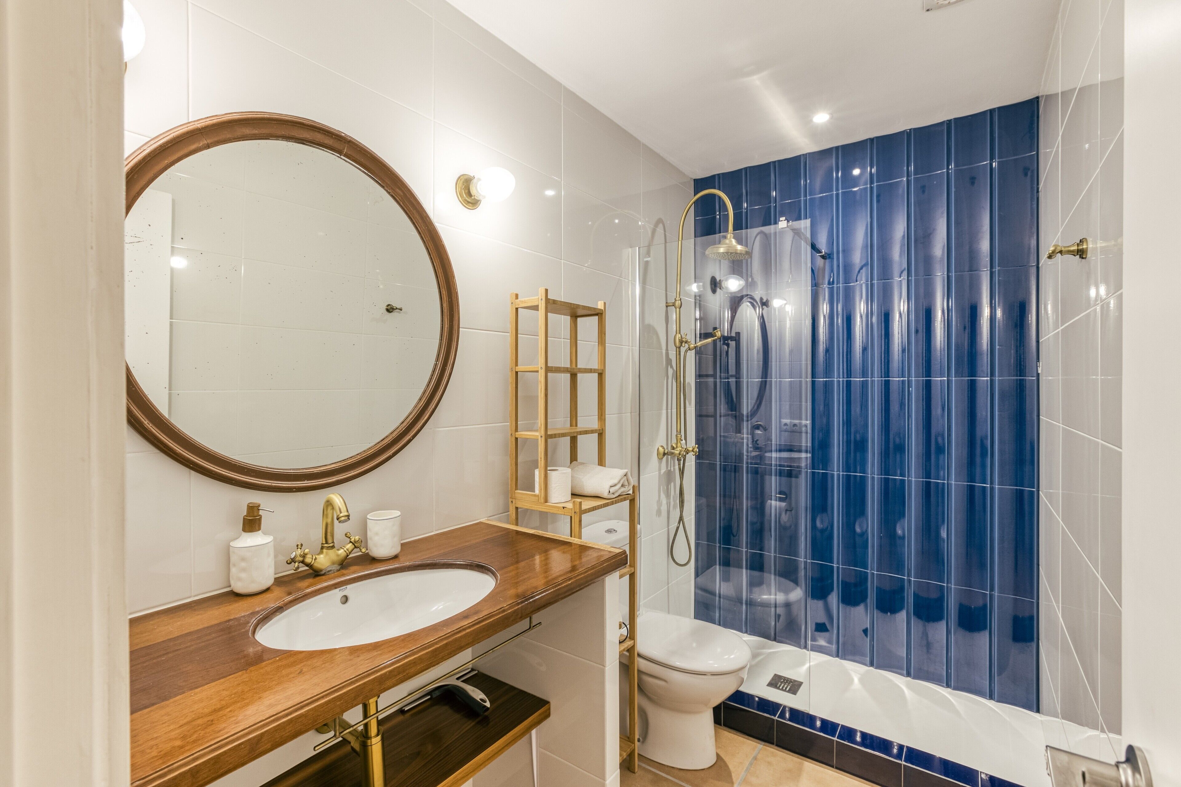 A bathroom with a wooden vanity, a large mirror, a toilet, and a shower with blue tiles.