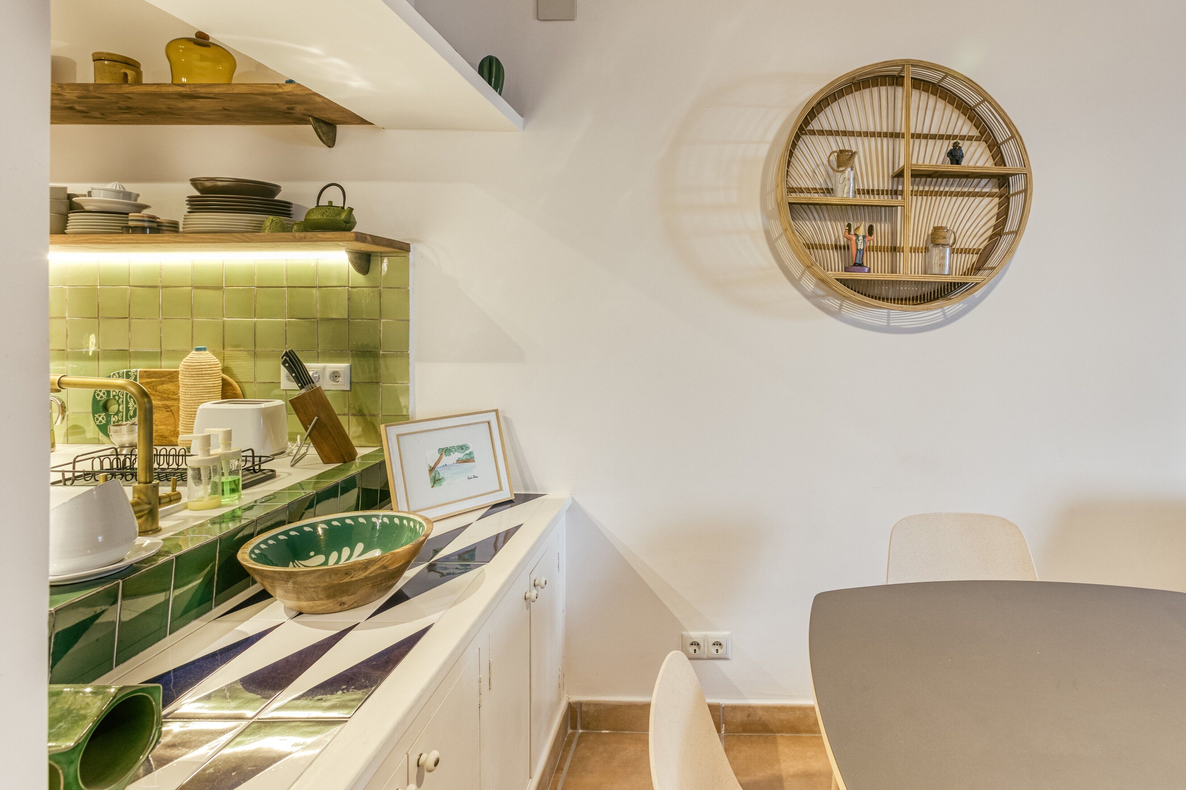 Een keuken met een groene betegelde achterwand, een houten plank met servies, een rond bamboe plankje en een witte stoel.