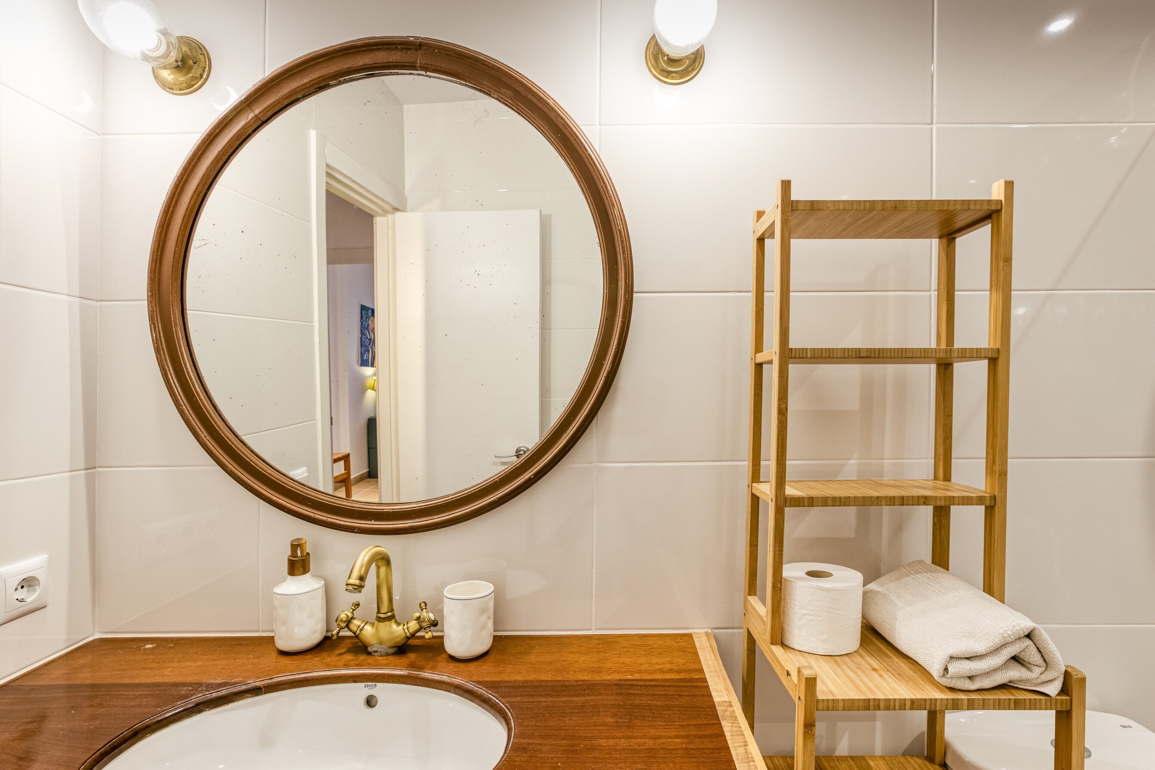 A bathroom with a wooden vanity, a round mirror, a wooden shelf with toiletries, and a white sink.