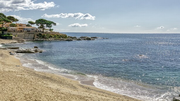 Una playa con aguas cristalinas, un litoral rocoso y edificios al fondo.