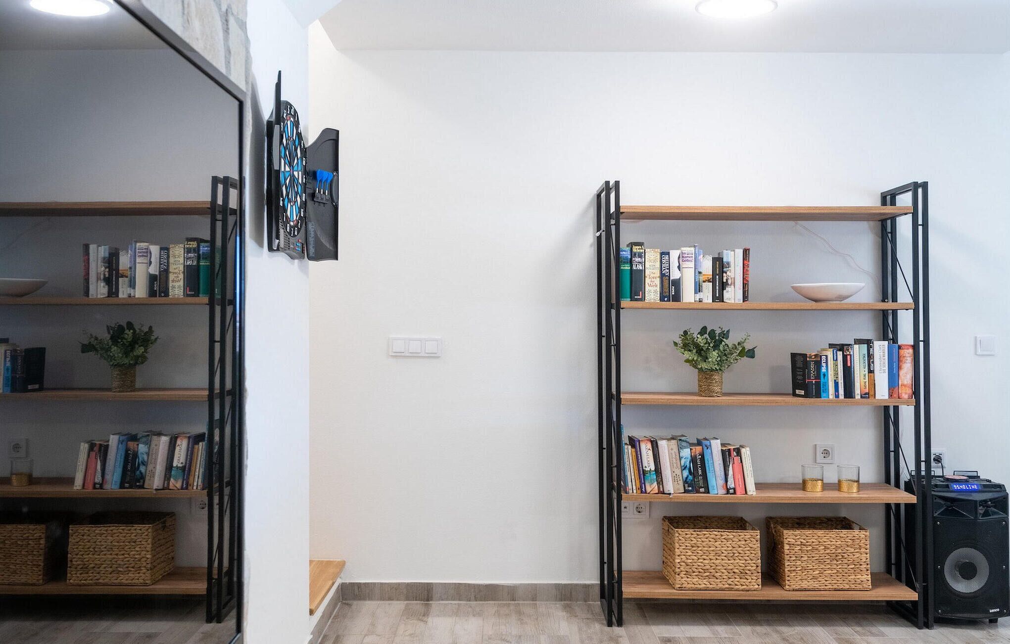 Une pièce avec deux étagères en métal noir adossées à un mur blanc, chaque étagère contenant des livres et de petites plantes. On trouve des paniers en osier sur les étagères du bas et un haut-parleur sur le côté droit.