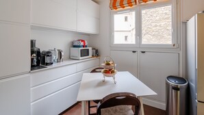 A compact kitchen with white cabinetry, a white table, a wicker fruit basket, a stainless steel trash bin, and a window with a striped valance. - Culture and Colour, by Veeve (Paris)