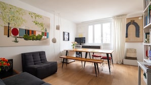 A modern living room with a sofa, a dining table with benches, and a kitchen area with a TV. - Culture and Colour, by Veeve (Paris)