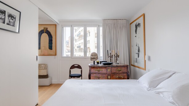 A bedroom with a large bed, a wooden dresser, a chair, and a window with curtains. - Culture and Colour, by Veeve (Paris)