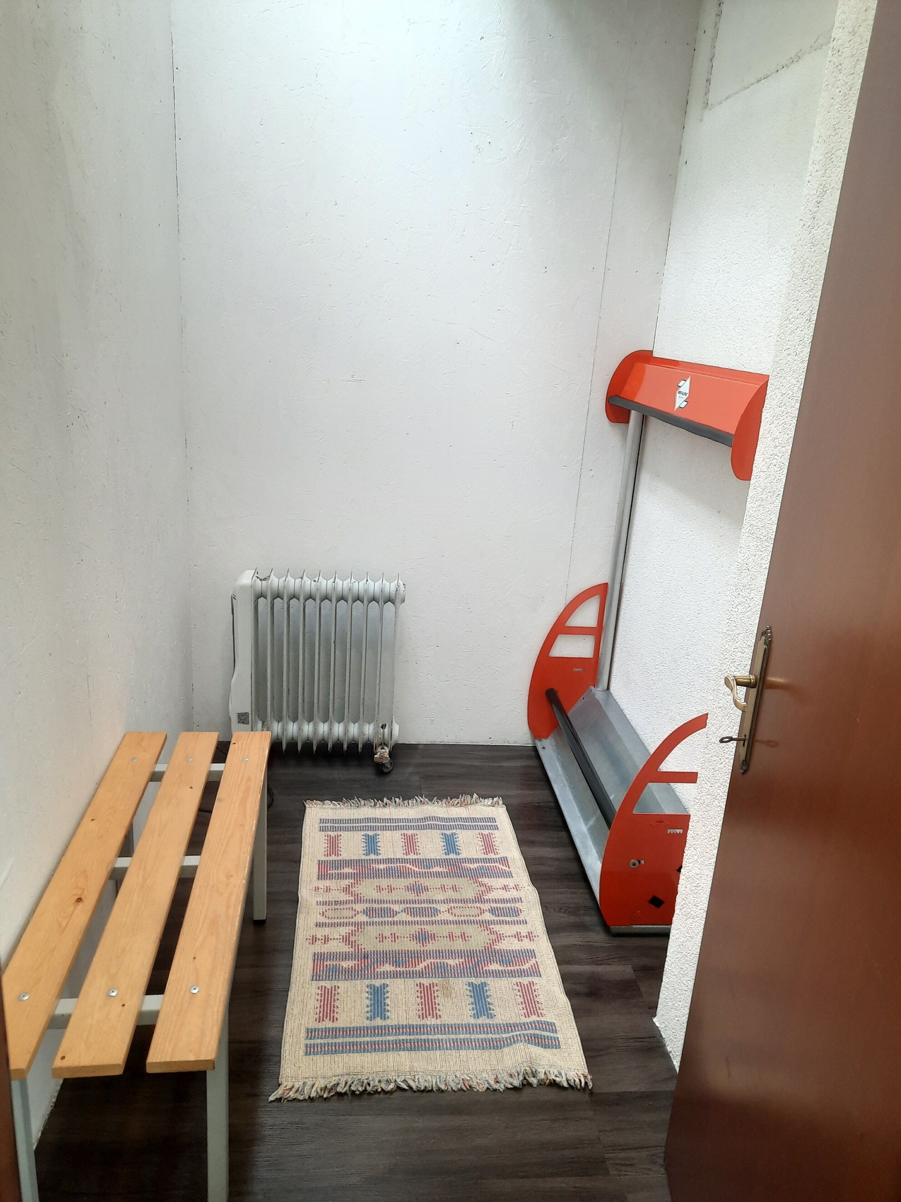 A small room with a wooden bench, a patterned rug, a radiator, and a red wall-mounted shelf.