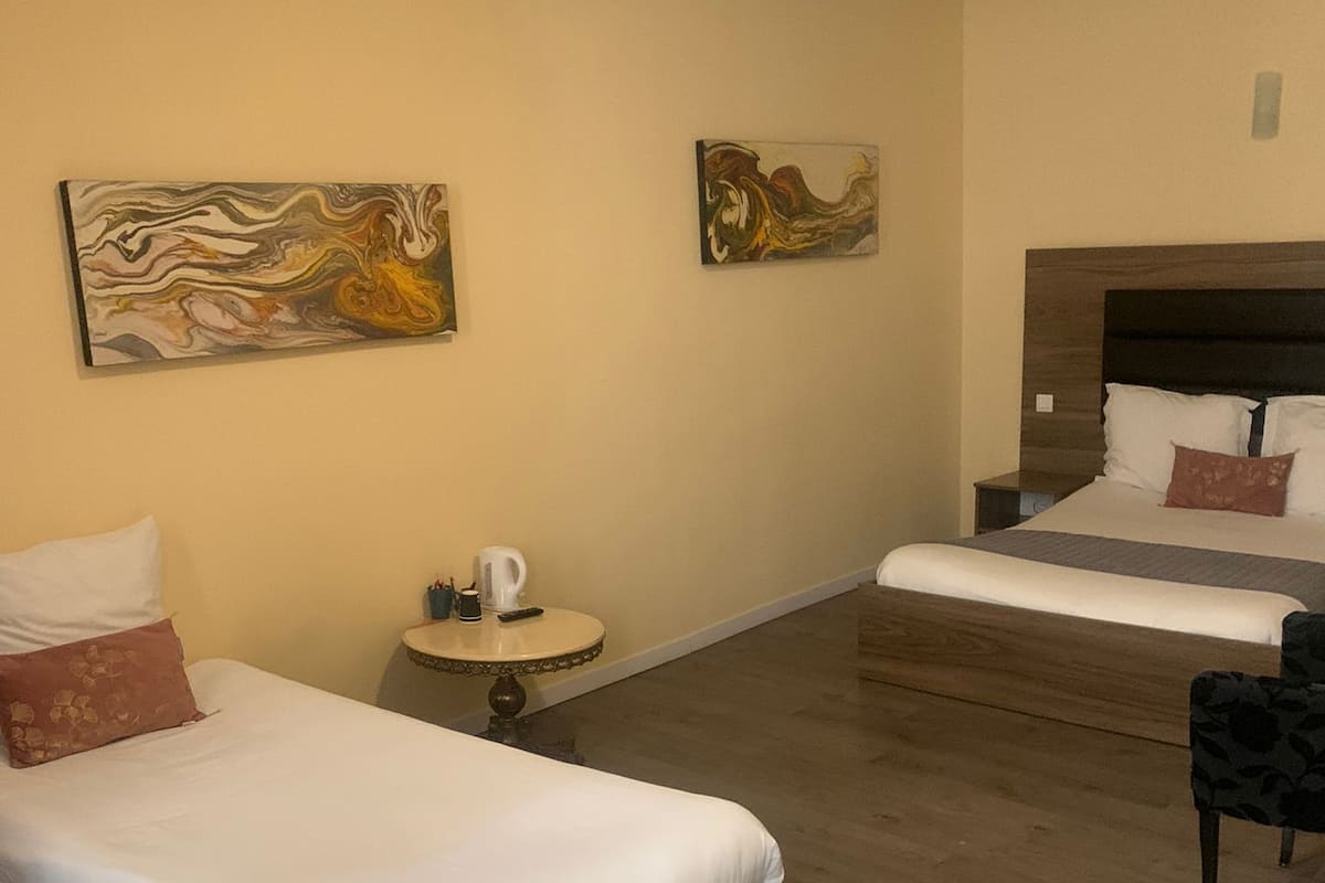 A hotel room with two beds, a small round table with a kettle, and two abstract paintings on the wall.