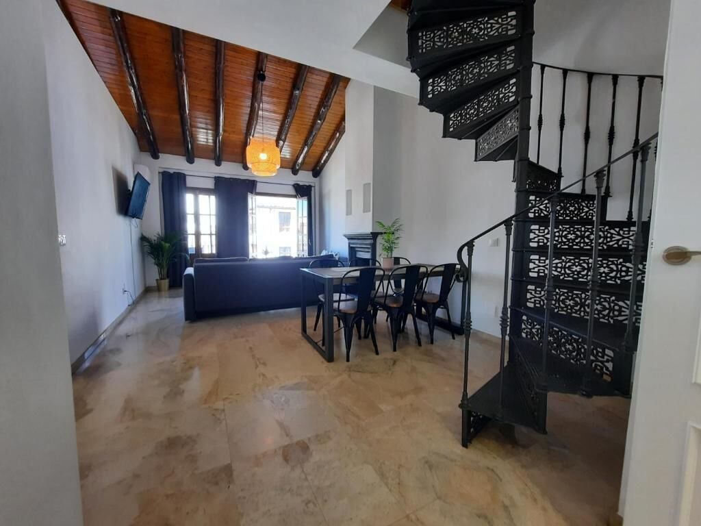 Amplia sala de estar con techo de madera, una escalera espiral negra y un comedor con mesa y sillas.