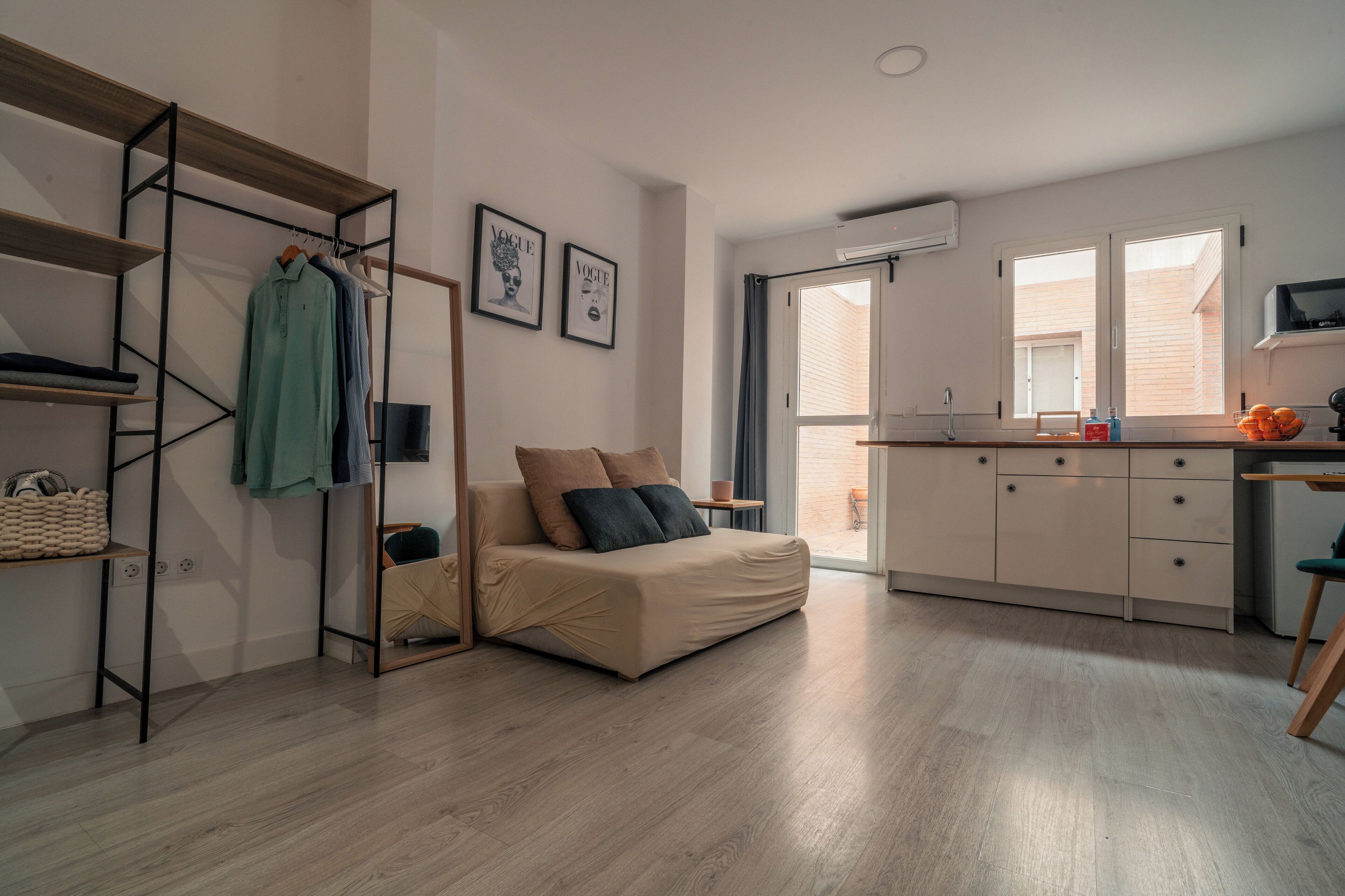 A compact living space with a sofa, a clothing rack, a small table, and a kitchen area.