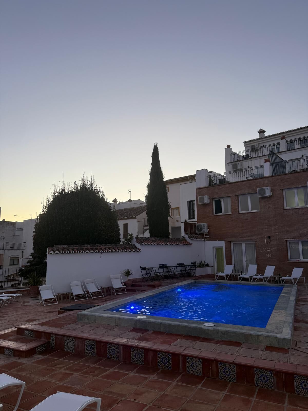 Ein Pool auf dem Dach mit Liegestühlen, eine geflieste Terrasse und ein Gebäude mit weißer Fassade und roten Ziegeldach.
