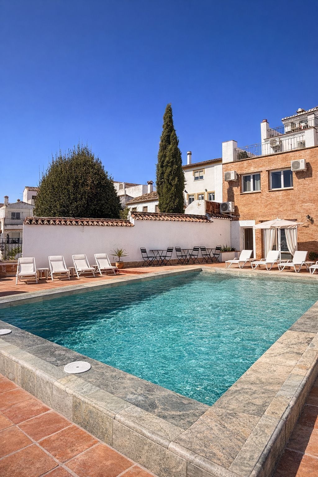 A rectangular swimming pool with clear blue water, surrounded by a stone deck. There are white lounge chairs aligned along the pool's edge.