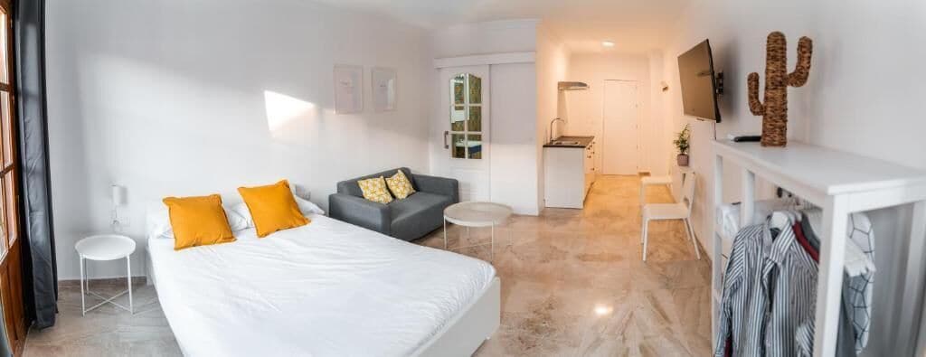 Una camera d'albergo moderna con un letto grande, un divano, un tavolino e una TV. Si nota una piccola cucina in fondo.