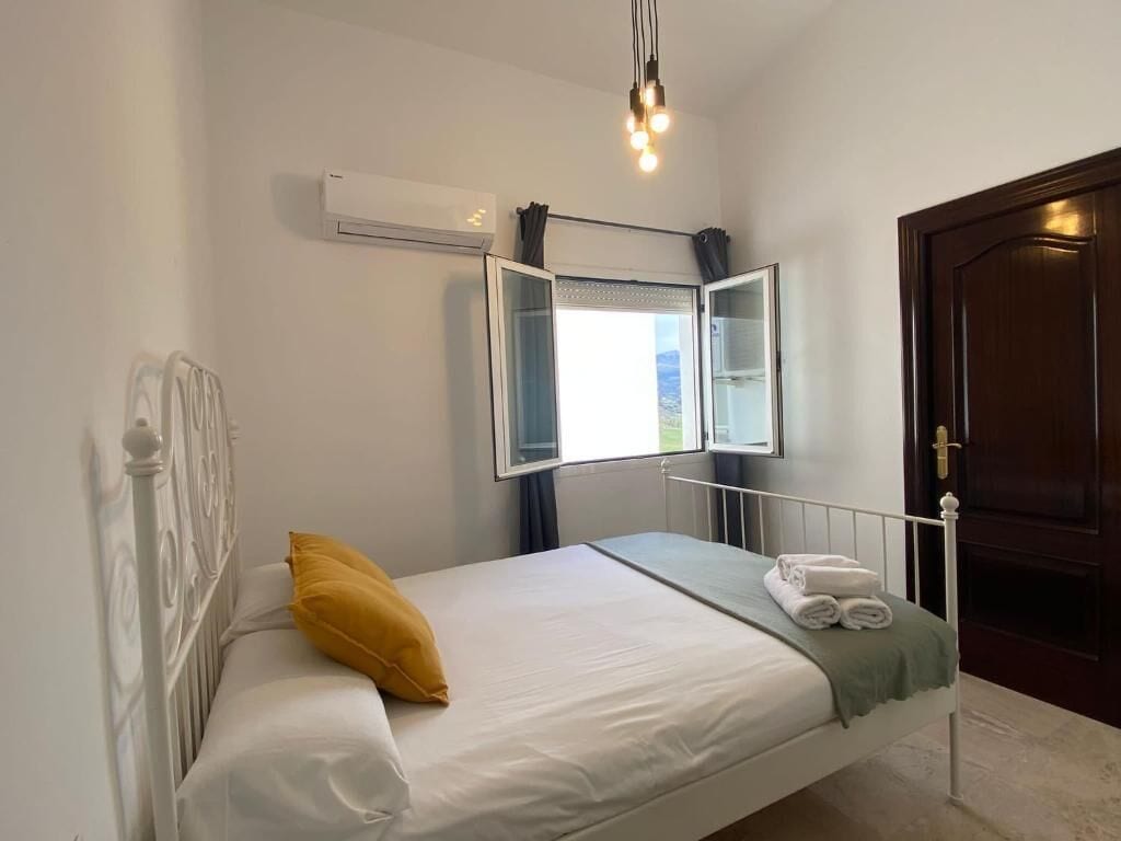 Una cama individual con ropa blanca, un almohábil amarillo y una toalla doblada. Se ve una ventana con cortinas, un equipo de aire acondicionado y una puerta con pomo de latón.