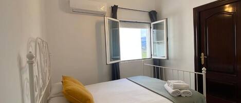 Una cama individual con ropa blanca, un almohábil amarillo y una toalla doblada. Se ve una ventana con cortinas, un equipo de aire acondicionado y una puerta con pomo de latón.