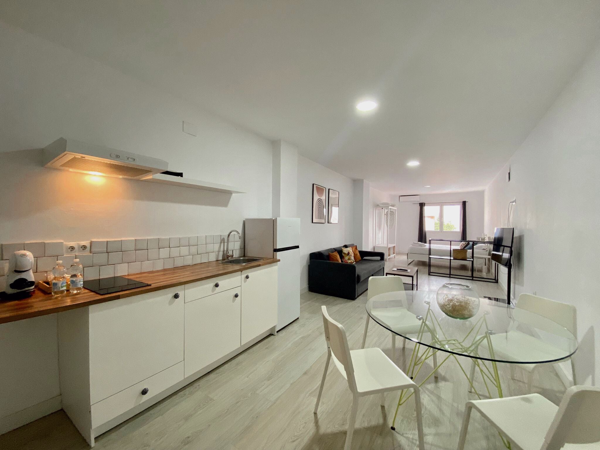 Una cocina moderna con armarios blancos, encimera de madera y estufa empotrada. Hay una zona de comedor con mesa de vidrio y sillas blancas.