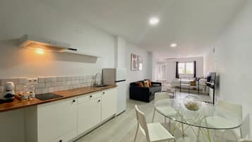 Una cocina moderna con armarios blancos, encimera de madera y estufa empotrada. Hay una zona de comedor con mesa de vidrio y sillas blancas.