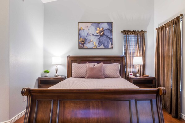 A neatly made bed with a headboard, two bedside tables with lamps, a wall-mounted painting, and a window with curtains.