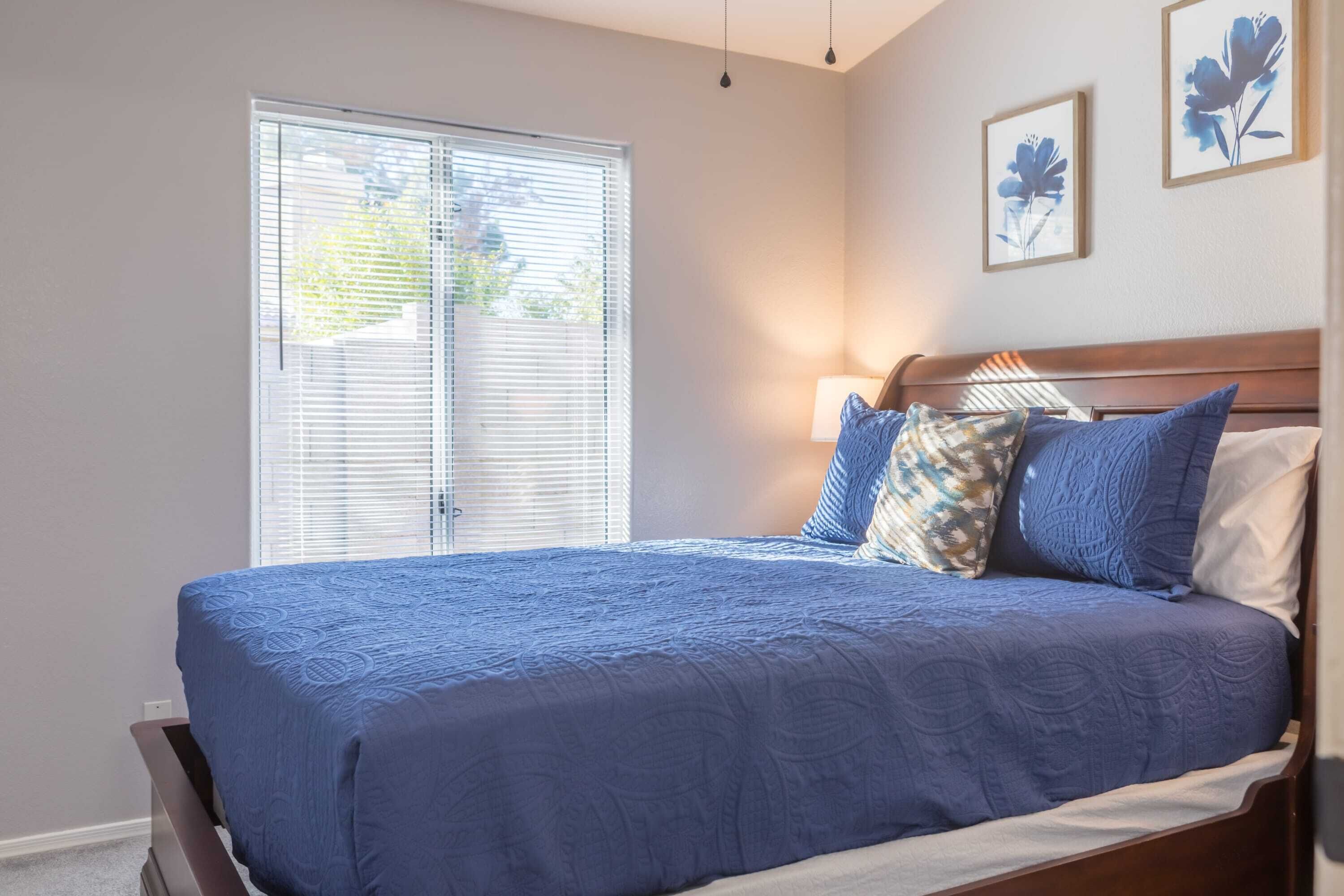 A bedroom with a large bed, a window with blinds, and two wall-mounted flower paintings.