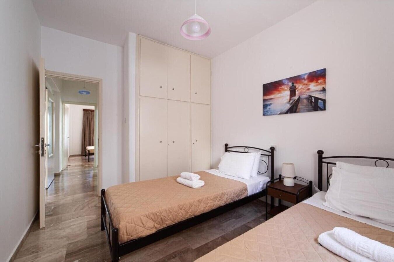 A bedroom with a bed, a bedside table, a wardrobe, a painting on the wall, and a hallway leading to another room.
