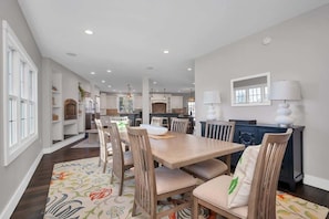 A spacious dining area with a long table and chairs, a kitchen in the background, and a fireplace with lamps. - Tee n’ Sea | Luxury Getaway - Golf & Coastal Views (Groton)