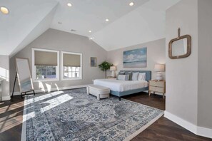 A bedroom with a large bed, a nightstand, a lamp, a painting, and a view of the outdoors. - Tee n’ Sea | Luxury Getaway - Golf & Coastal Views (Groton)