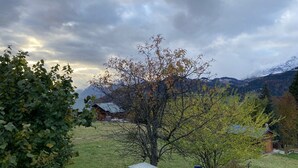 Ländliche Landschaft mit freiem Blick auf ein Gebirge im Hintergrund, einem Baum im Vordergrund und einem kleinen Gebäude mit geneigtem Dach.