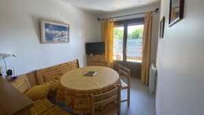 Ein Essbereich mit einem runden Tisch, Holzstühlen und einer Couch. Ein Fernseher steht auf einem Schrank, an der Wand hängt ein Bild und ein Fenster ist mit Vorhängen drapiert.