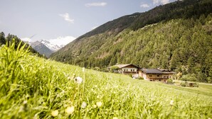 Une maison avec un toit en pente, entourée d’un champ verdoyant et de montagnes boisées.