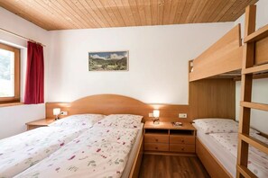 A bedroom with a wooden bunk bed, a single bed with floral bedding, a nightstand with a lamp, a wall-mounted picture, and a window with red curtains.