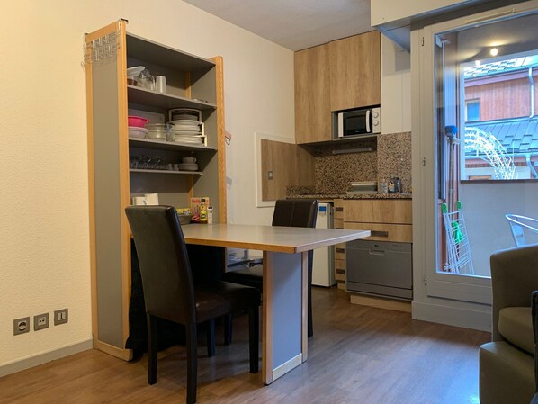 Une cuisine compacte avec des meubles en bois, un four à micro-ondes et un lave-vaisselle. On y trouve une table à manger avec des chaises, ainsi qu’une petite étagère avec de la vaisselle et des verres.