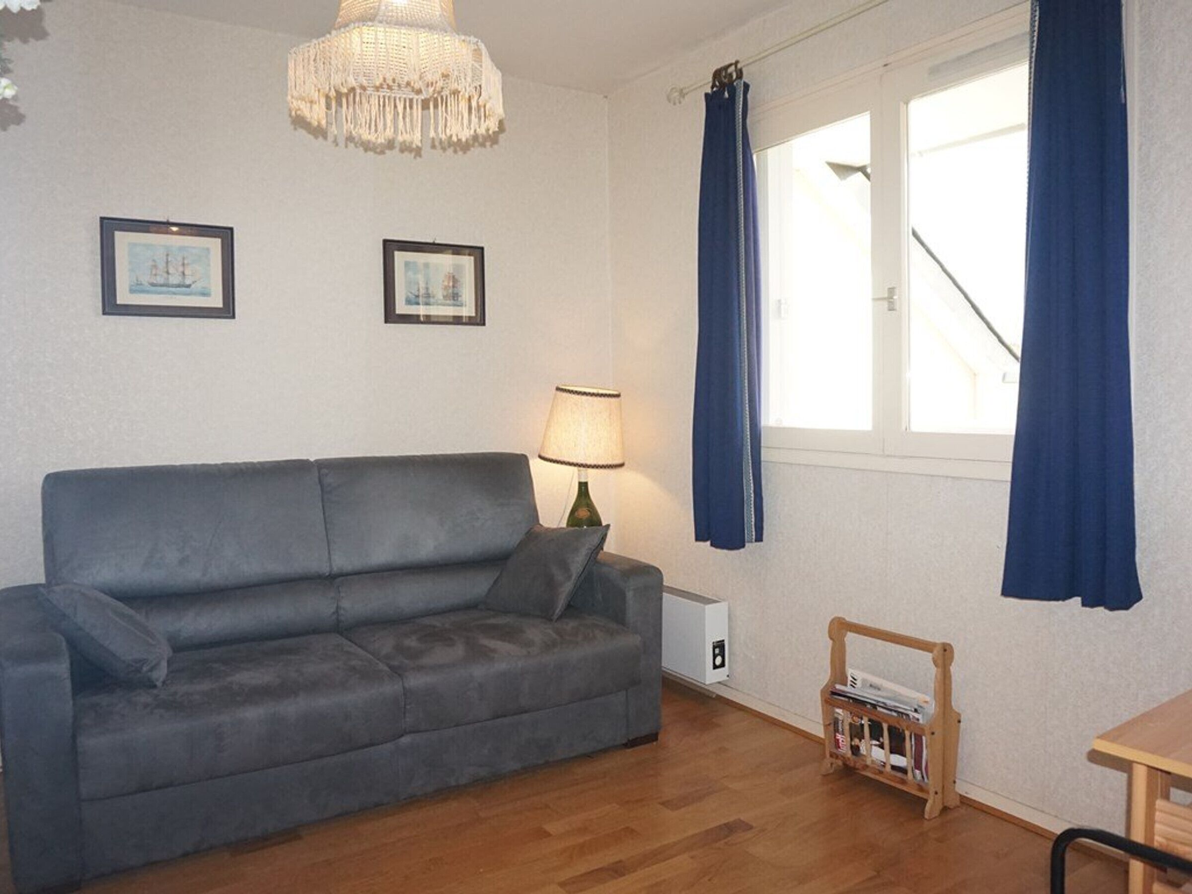 A room with a grey sofa, a small table, a lamp, a bookshelf, and a window with curtains.