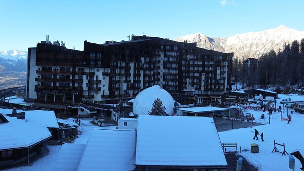 Een hotel met meerdere verdiepingen en balkons, met een skigebied op de achtergrond en een besneeuwde omgeving.