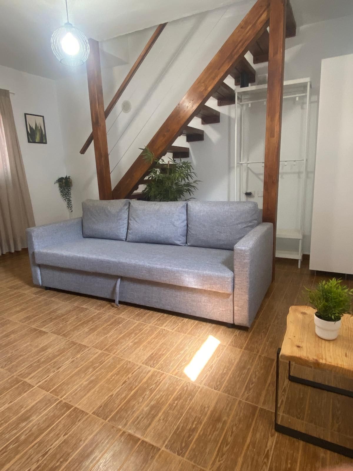 Una sala de estar con sofá gris, piso de madera, una escalera con vigas a la vista y una planta en maceta.