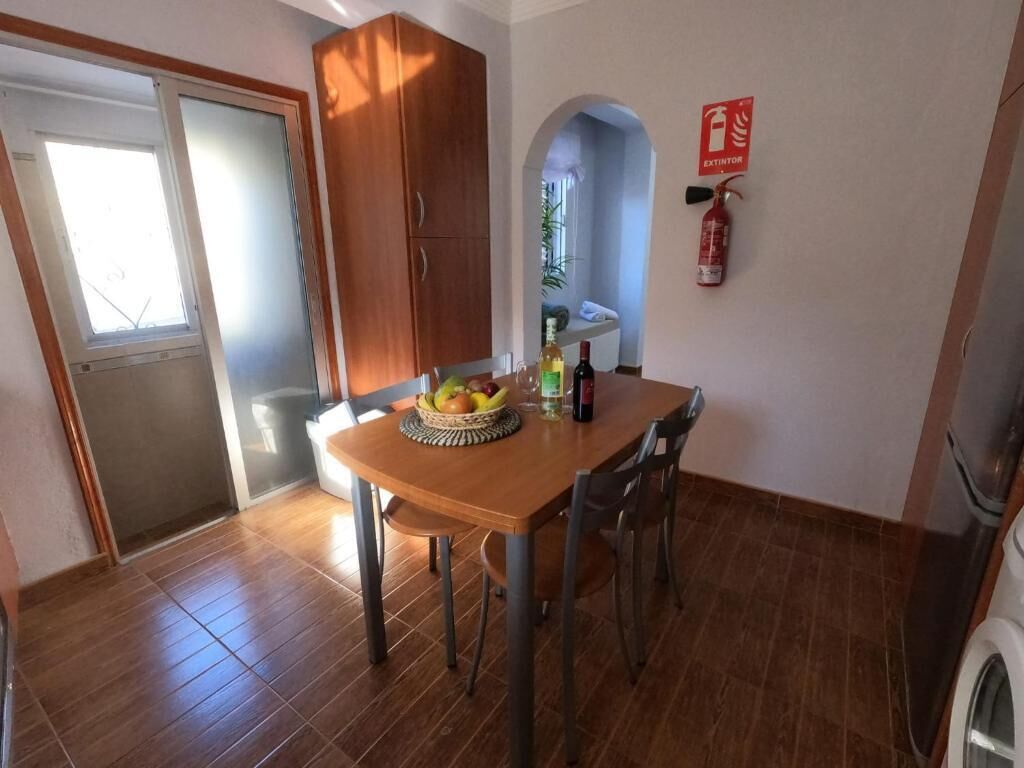 A dining area with a wooden table, chairs, and a fruit bowl. There is a fire extinguisher on the wall and a washing machine in the corner.