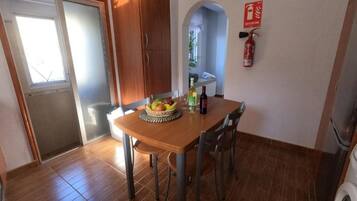 A dining area with a wooden table, chairs, and a fruit bowl. There is a fire extinguisher on the wall and a washing machine in the corner.