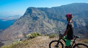 A person standing next to a mountain bike overlooking a mountainous landscape with a clear view of the valley below. - Alojamientos Charco Azul (Agaete)