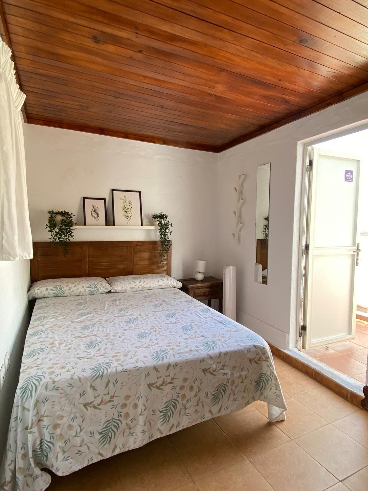 Une chambre avec un plafond en bois, un lit recouvert d’une couvre-lit à motifs floraux, une table de chevet, un miroir fixé au mur et une porte ornée du symbole du Wi-Fi.